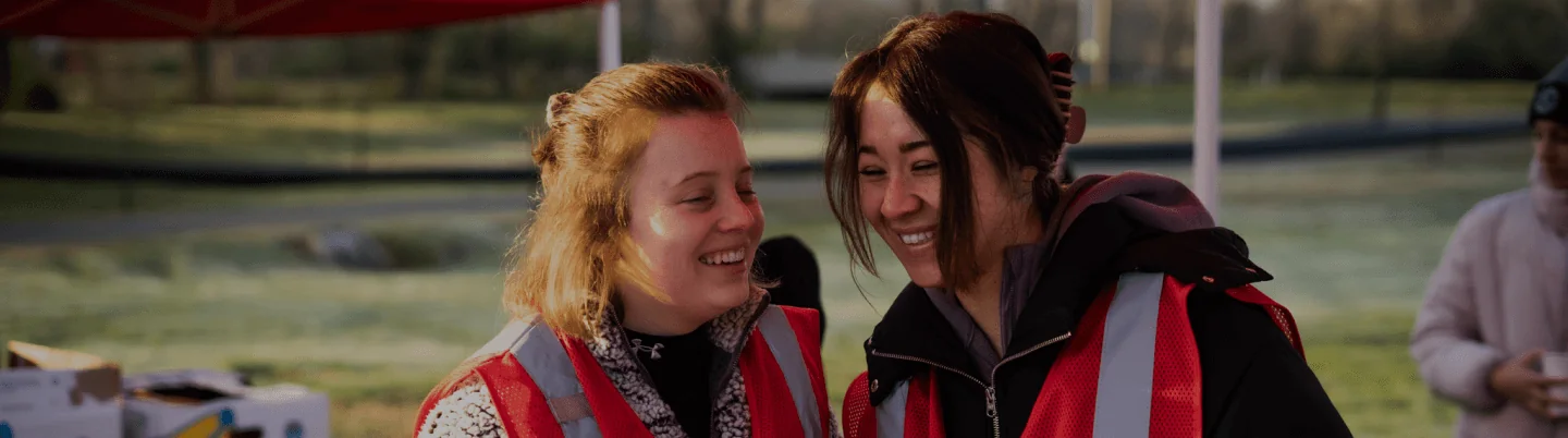 Two girls laughing with each other outside during a conversation wearing red safety vests.